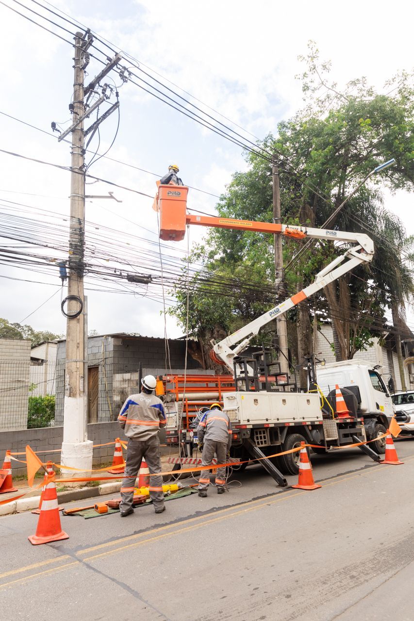 Equipe de manutenção de estradas, vestindo equipamentos de segurança laranja, repara cabos de energia a partir de um caminhão com plataforma elevatória ao lado de uma rua.