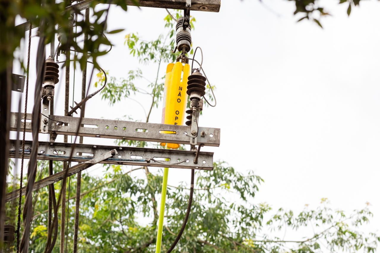 Poste de energia danificado com isoladores e fios contra um céu frondoso.