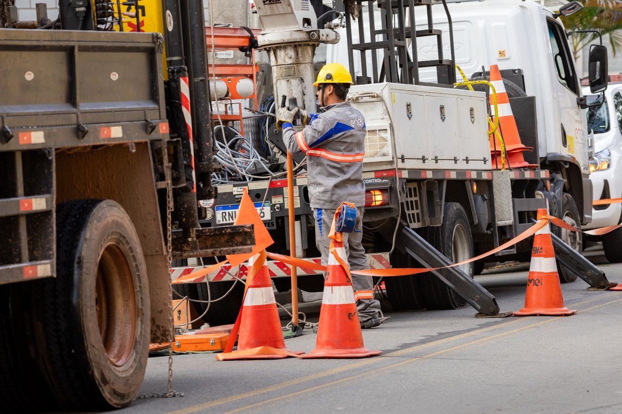 Equipe de obras rodoviárias com cones laranja e caminhão de construção bloqueando uma rua enquanto um operário opera um equipamento.
