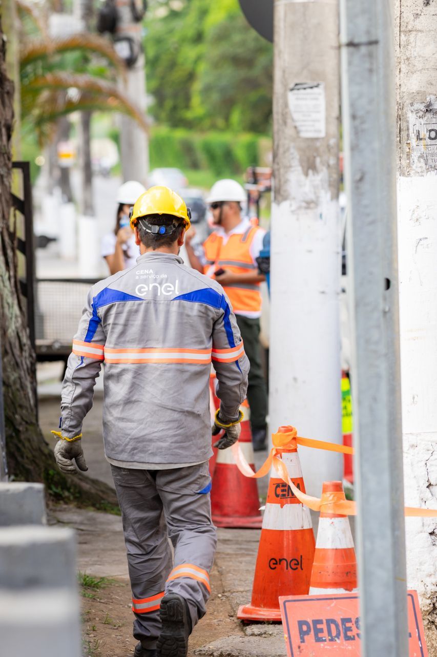 Operários da construção civil com capacetes caminham ao lado de cones de segurança laranja em uma calçada da cidade.
