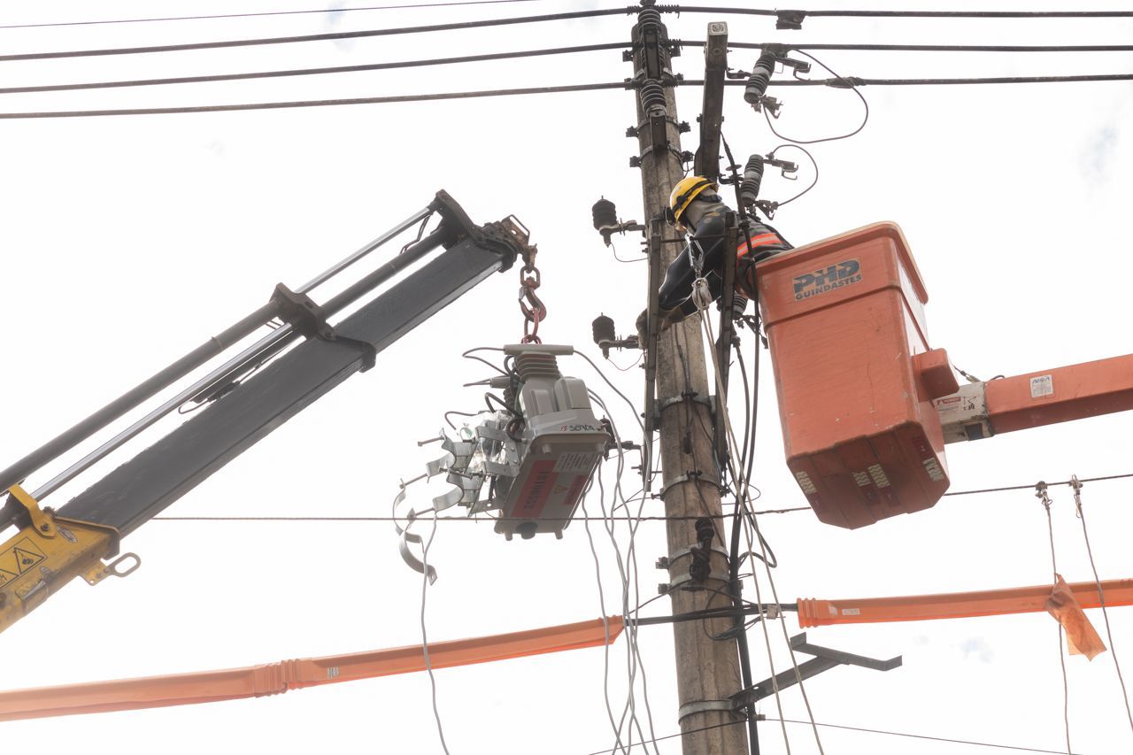 Dois caminhões com cestos elevatórios trabalham em linhas de energia ao lado de um poste com fios emaranhados.