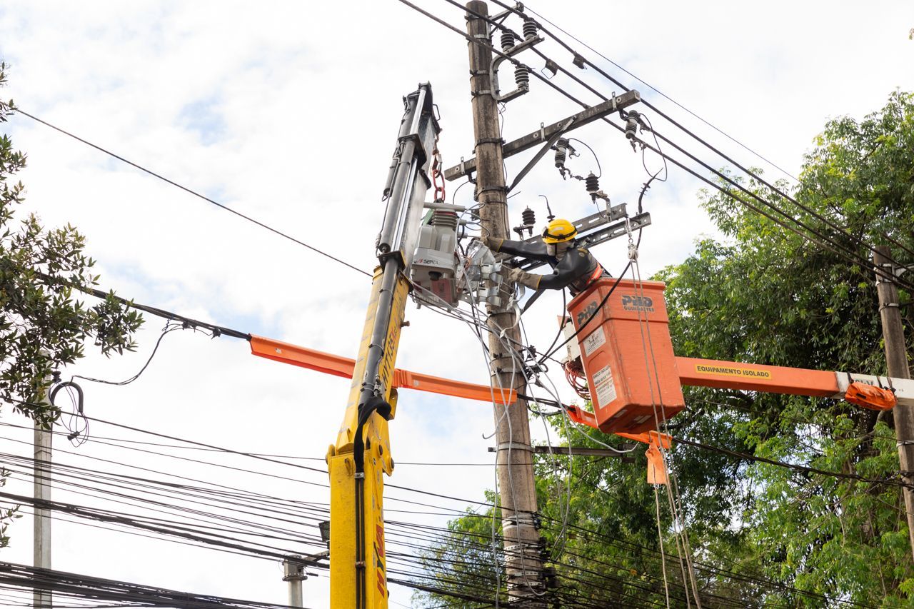 Operários de serviços públicos, vestidos com plataformas elevatórias laranja, reparam cabos de energia em um poste acima da rua.