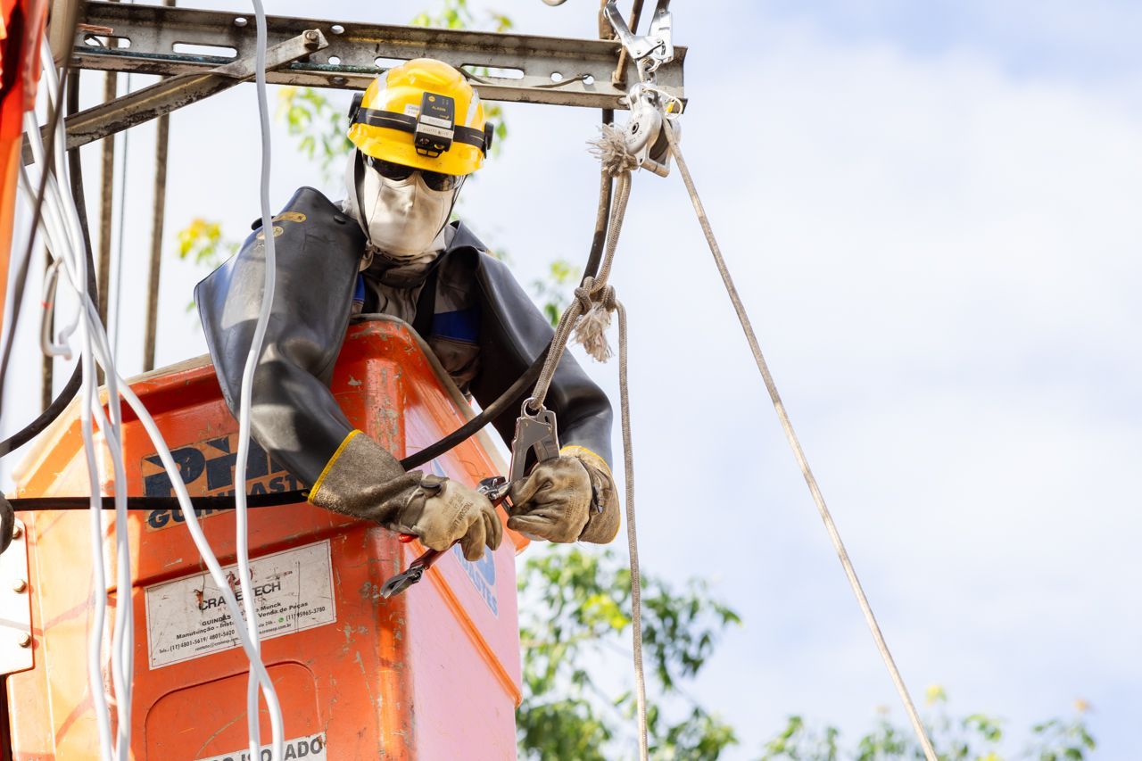 Operário com capacete amarelo e equipamento de segurança em pé em uma plataforma elevatória laranja, manuseando cabos a grande altura.