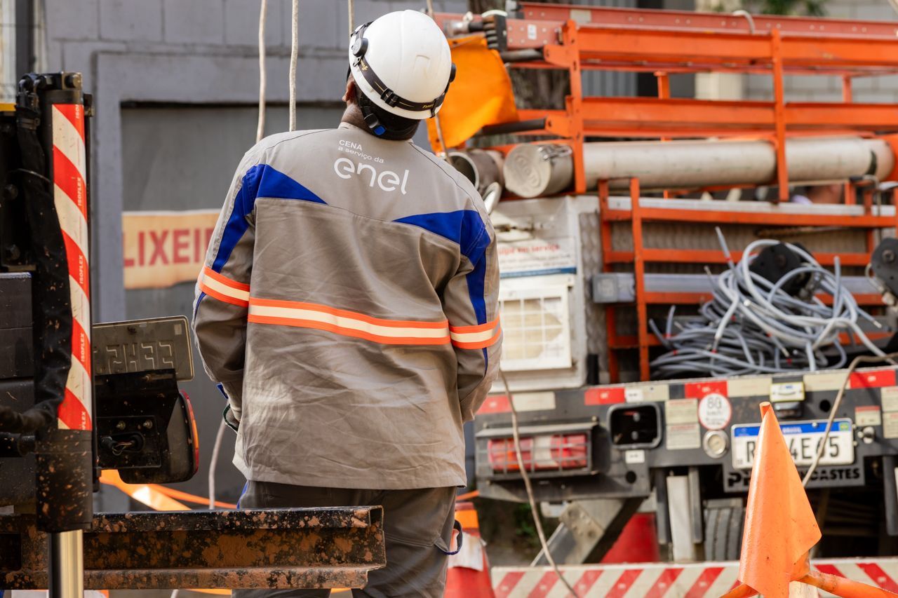 Operário da construção civil com capacete, em pé perto de canos e equipamentos laranja em um canteiro de obras.