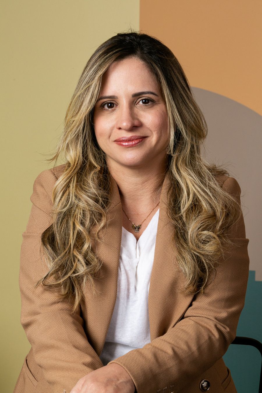 Mulher sentada contra um fundo bege, vestindo um blazer bege e uma blusa branca, sorrindo para a câmera.
