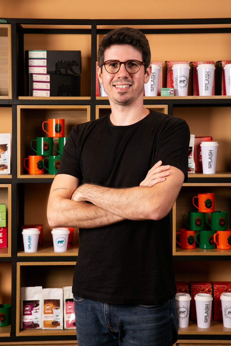Homem em pé com os braços cruzados em frente a prateleiras com caixas e copos de produtos coloridos.