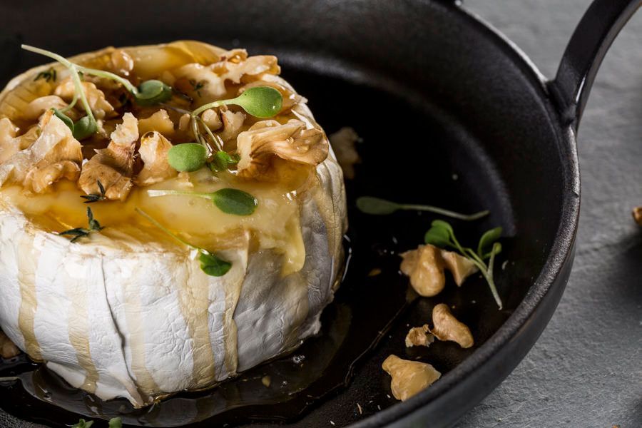 Queijo em formato de roda assado em frigideira preta, coberto com mel, nozes e ervas.