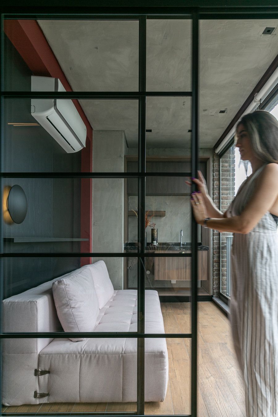 Mulher em pé junto a uma divisória de vidro em uma sala de estar moderna com sofá branco e ar-condicionado de parede.