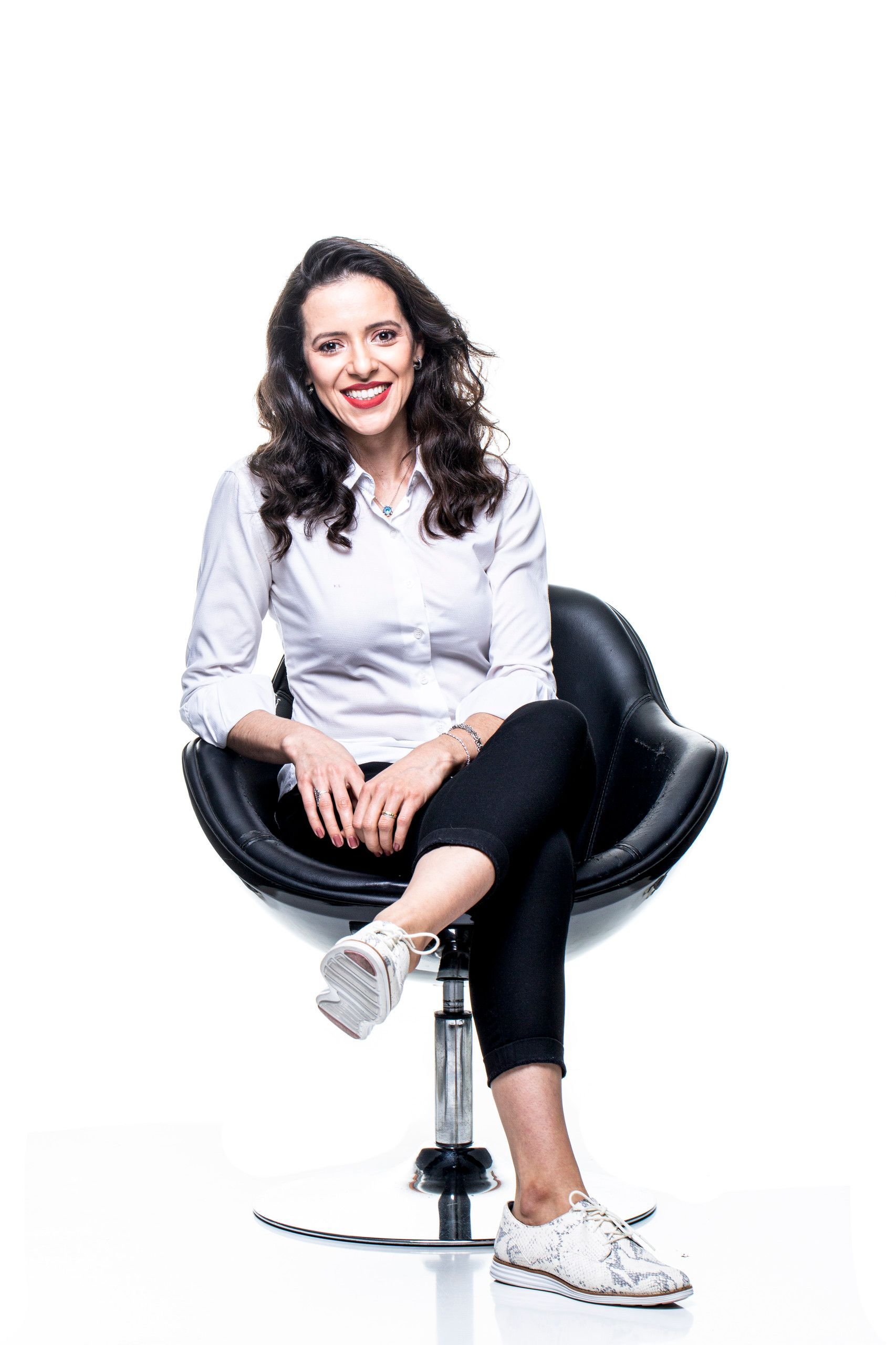 Mulher sentada em uma cadeira preta, sorrindo, vestindo uma blusa branca, calça escura e tênis brancos.