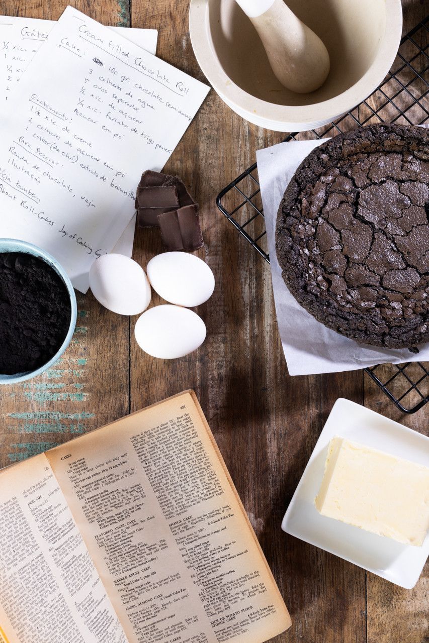 Ingredientes e utensílios para bolo sobre uma mesa de madeira, incluindo bolo de chocolate, ovos, farinha, manteiga e anotações da receita.