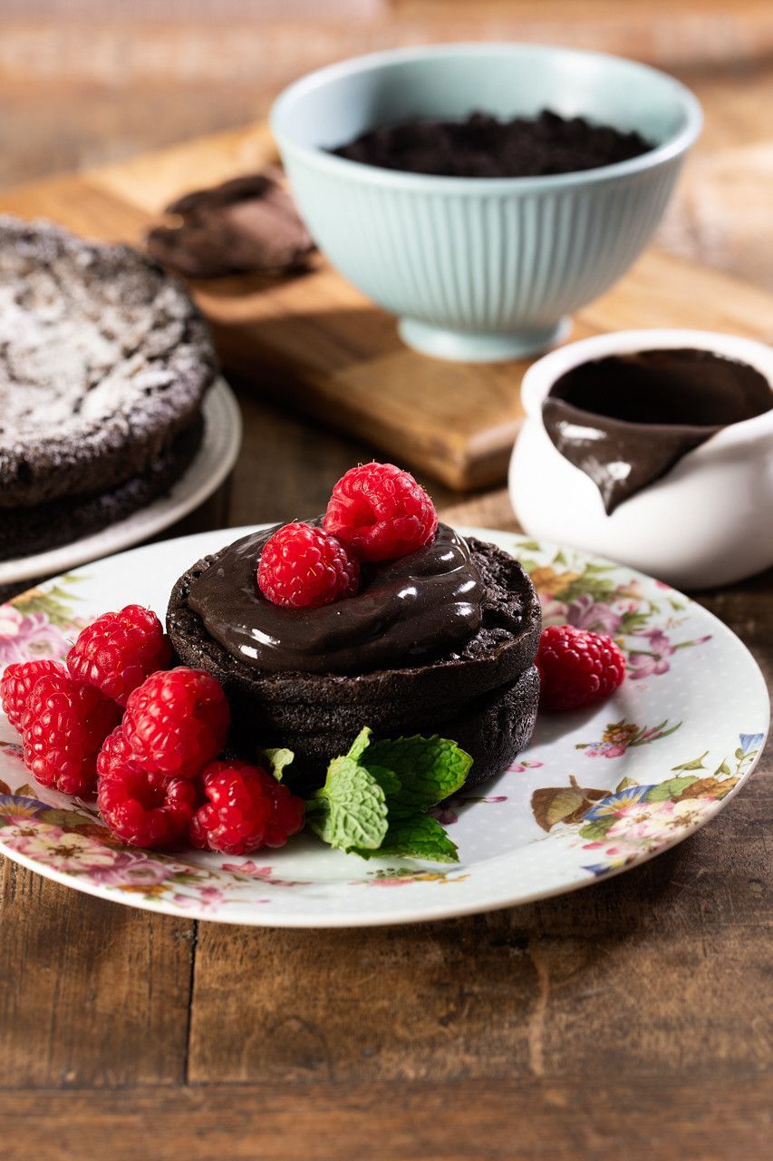 Bolos de chocolate e framboesas sobre uma mesa rústica, com calda de chocolate em uma tigela e xícara brancas.