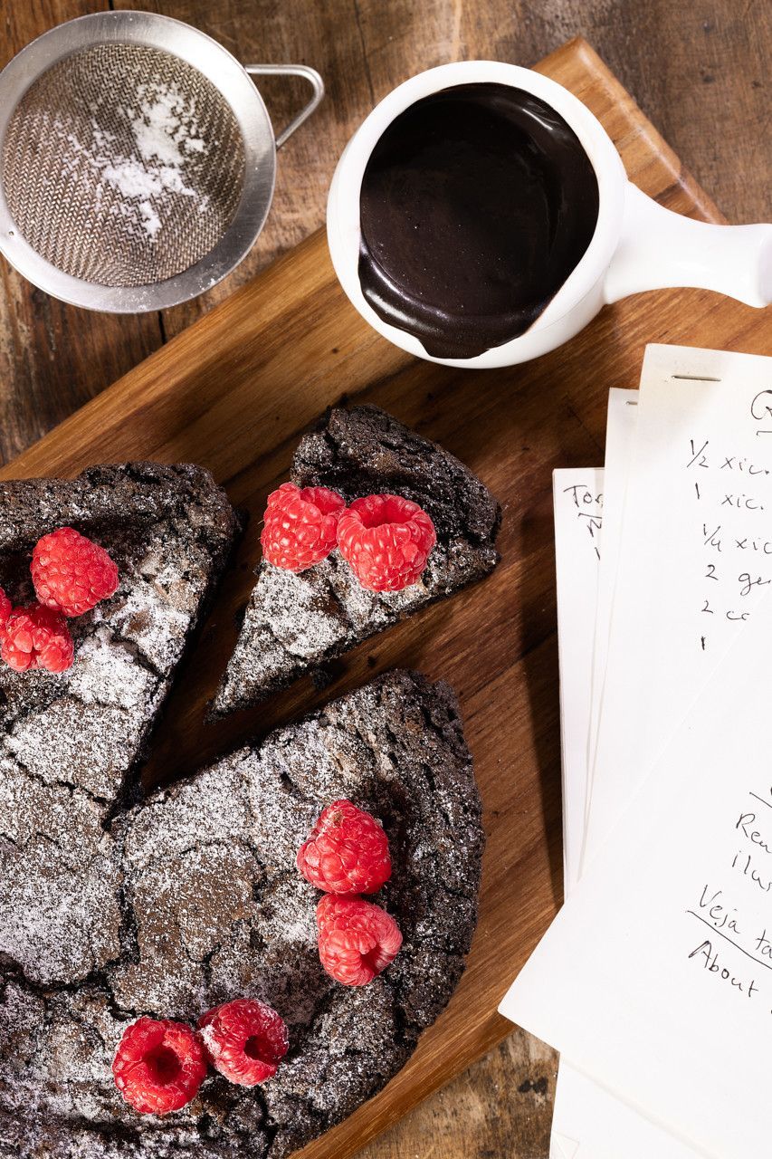 Fatias de bolo de chocolate com framboesas em uma tábua de madeira ao lado de uma xícara de café e uma peneira.