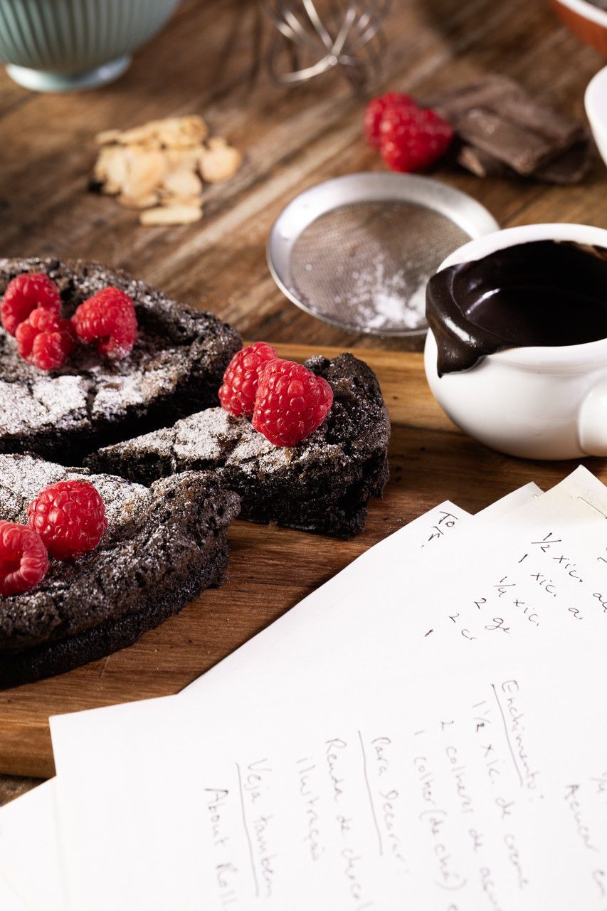 Brownies de chocolate com framboesas e açúcar de confeiteiro ao lado de um caderno e calda de chocolate em uma mesa de madeira.
