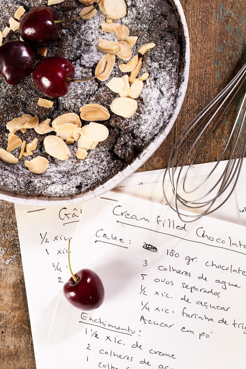 Bloco de notas com receita escrita à mão ao lado de uma tigela de frutas vermelhas, amêndoas fatiadas e um batedor de arame sobre uma superfície de madeira.