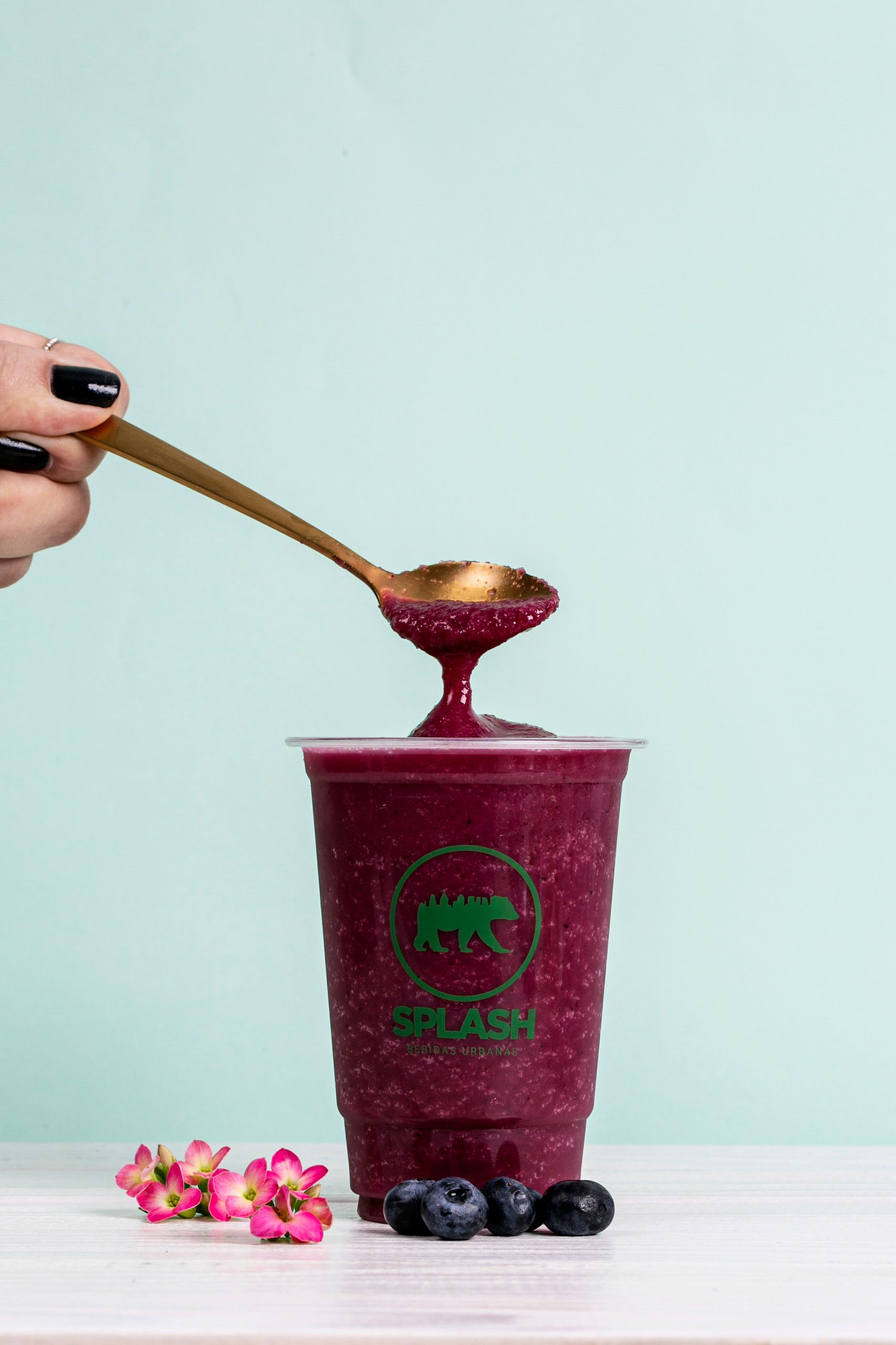 Uma pessoa serve manualmente uma bebida rosa com uma colher para um copo cor de vinho com frutas vermelhas sobre uma mesa branca.
