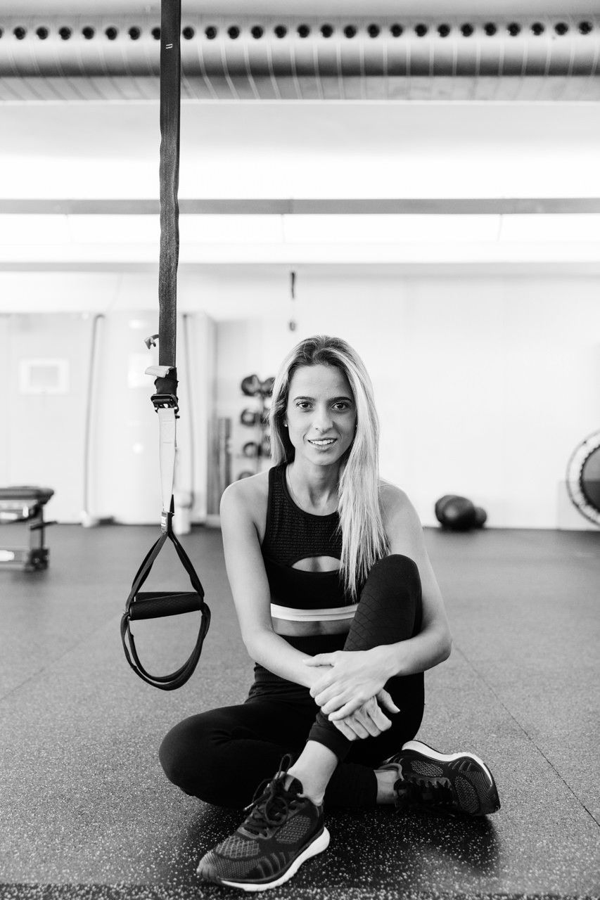 Mulher sentada no chão da academia ao lado de tiras de suspensão penduradas, sorrindo em um espaço para exercícios.