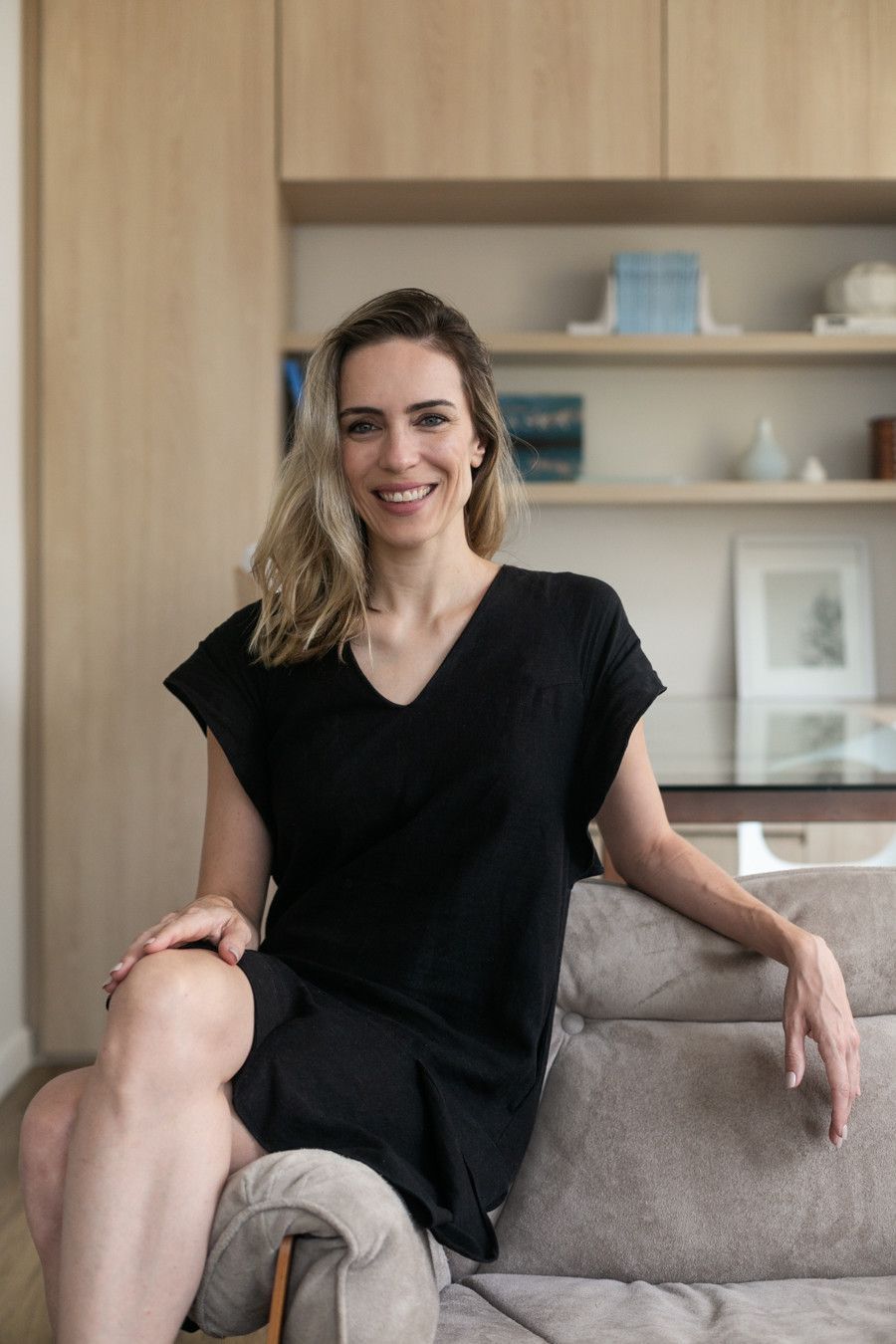 Mulher sentada em um sofá cinza em uma sala de estar moderna, sorrindo e vestindo um vestido preto.