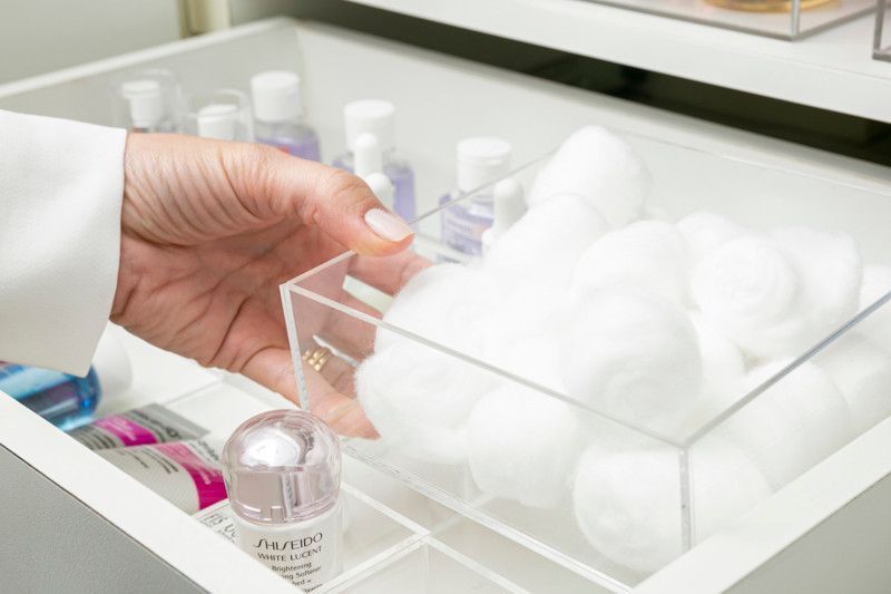 Mão alcançando um recipiente transparente com bolas de algodão branco em uma gaveta do banheiro.