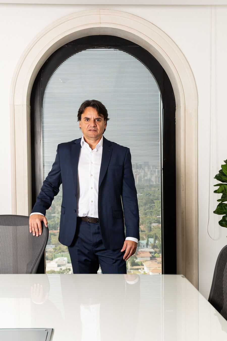 Um homem de terno azul-marinho está de pé ao lado de uma mesa de conferência branca em uma porta arqueada com persianas atrás dele.