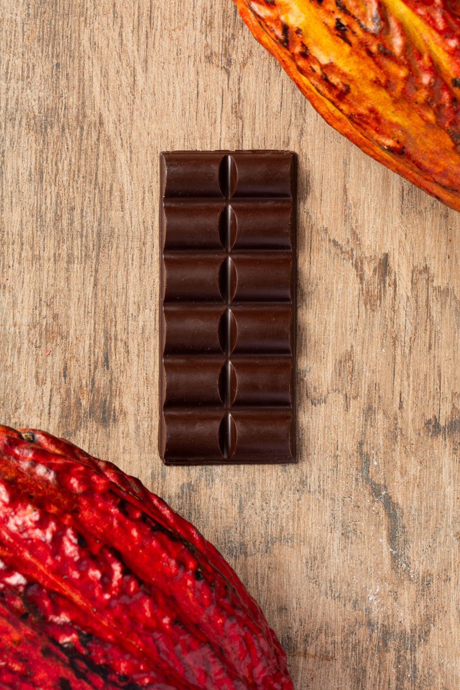 Barra de chocolate amargo sobre uma superfície de madeira, emoldurada por vagens de cacau vermelhas e laranjas.