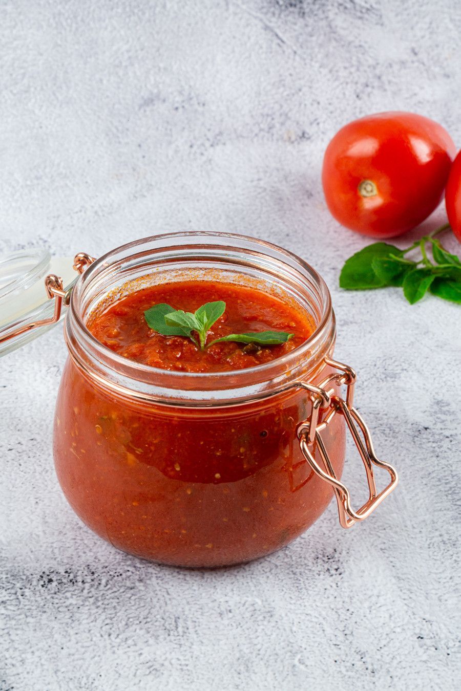 Pote de molho de tomate com manjericão para decorar e tomates frescos sobre uma superfície leve.