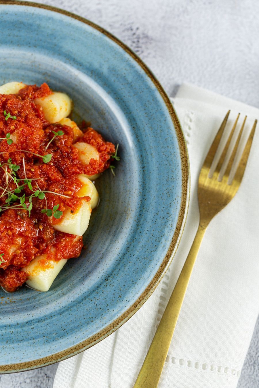 Prato azul de massa com molho de tomate vermelho e queijo ralado sobre um guardanapo branco, com um garfo dourado ao lado.