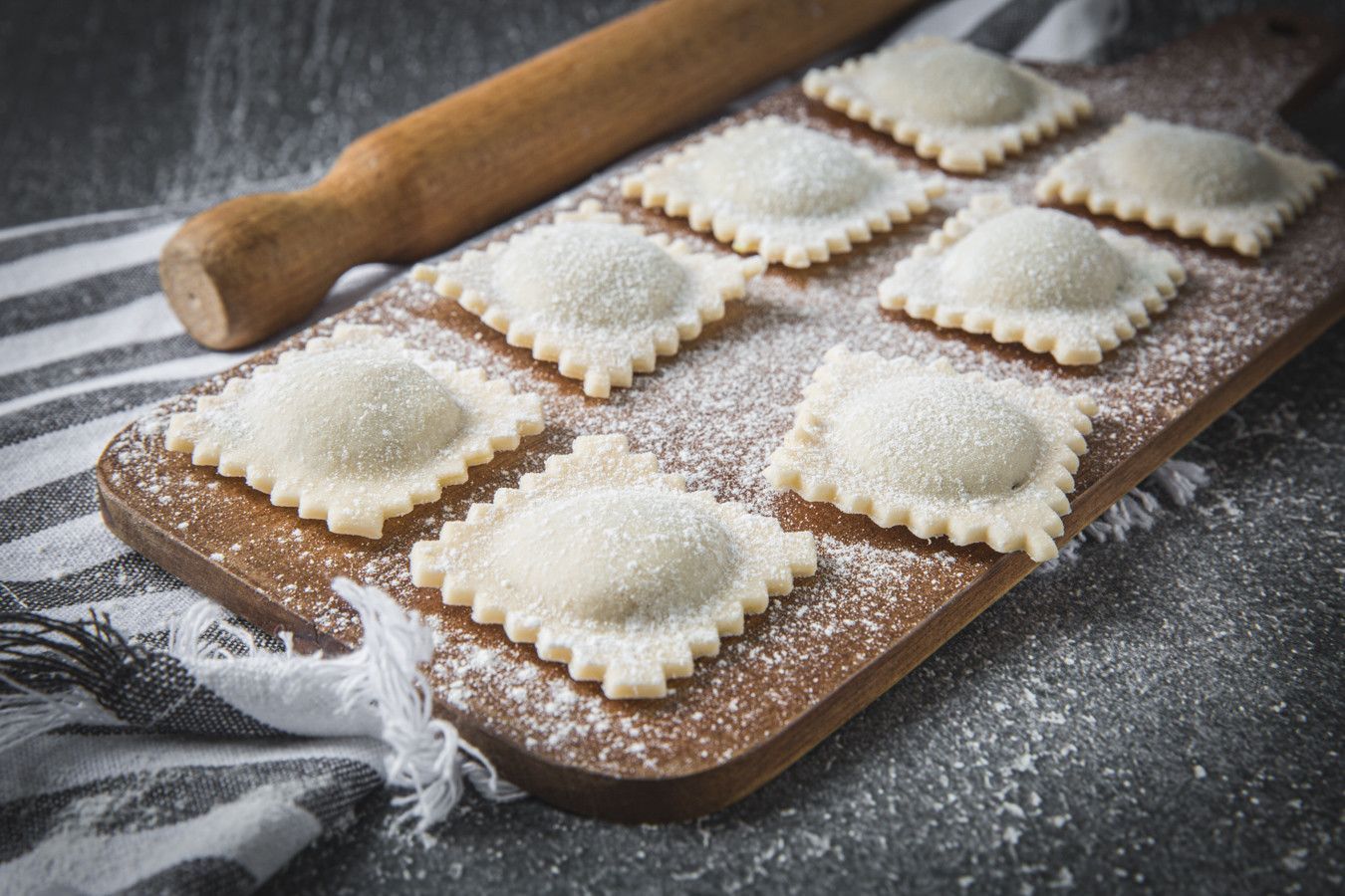 Quadrados de massa de ravioli polvilhados com farinha sobre uma tábua de madeira, com um rolo de massa por perto.