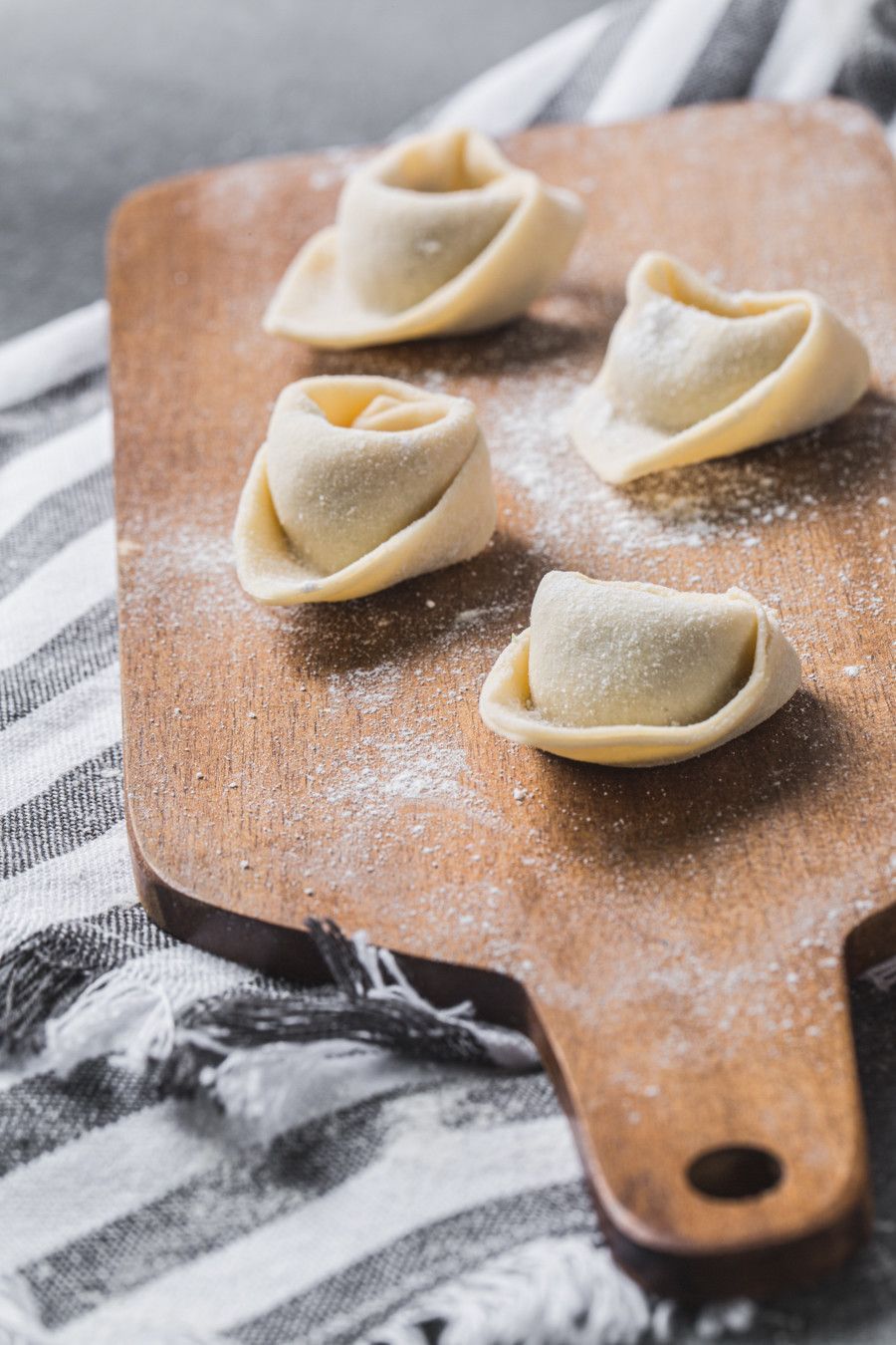 Quatro tortellini crus sobre uma tábua de madeira enfarinhada com um pano listrado ao fundo.