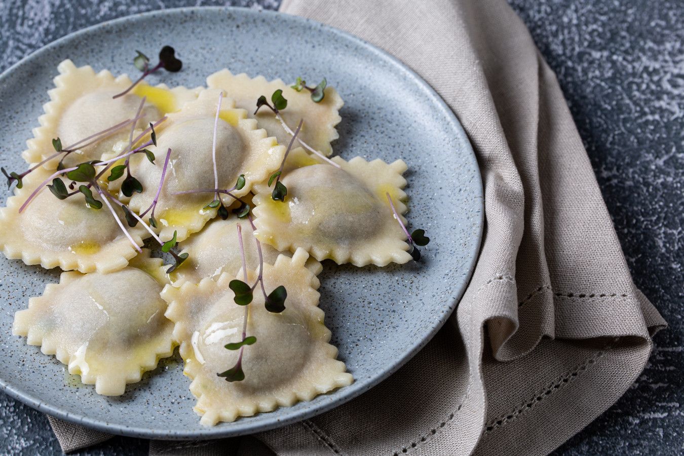 Ravioli num prato azul com microverdes, ao lado de um guardanapo de linho dobrado sobre uma mesa escura.