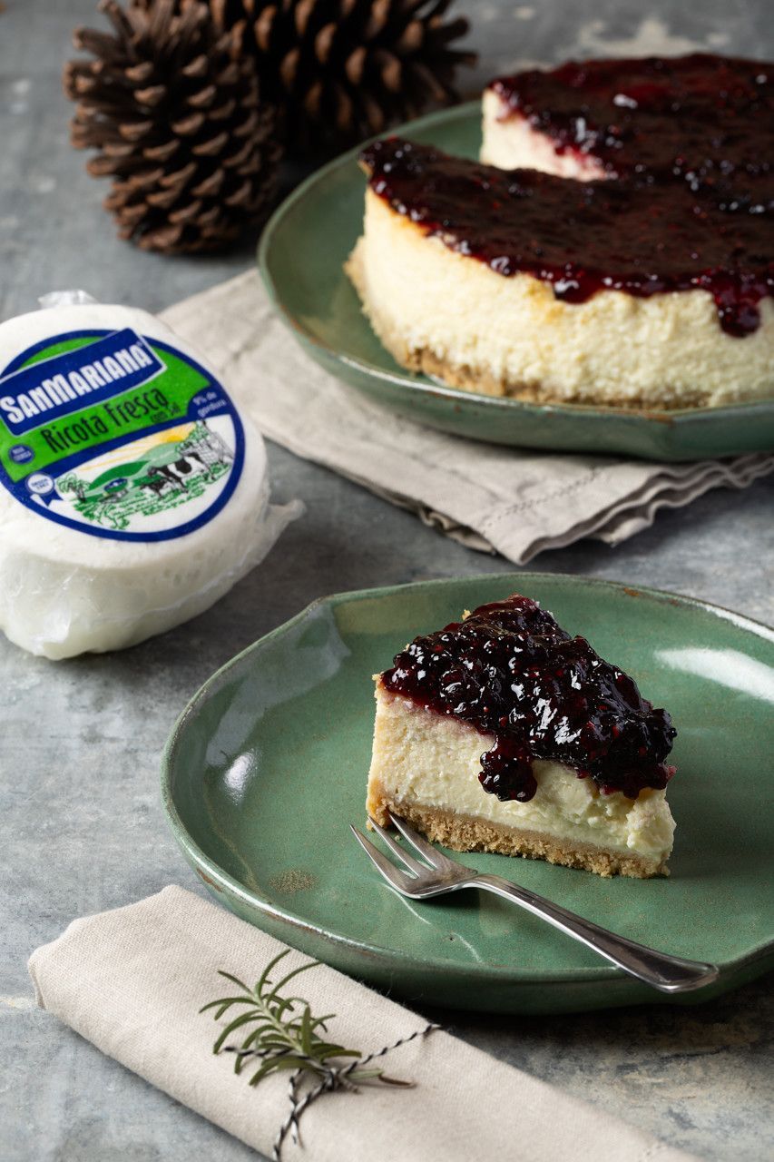 Fatias de cheesecake cobertas com calda de frutas vermelhas em pratos verdes ao lado de um pote de queijo ricota.