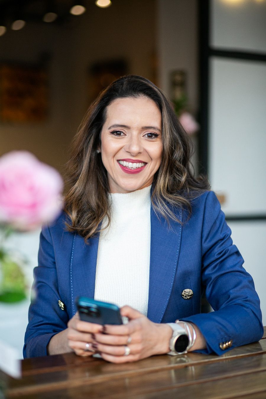 Mulher sorridente de blazer azul segurando um telefone em uma mesa de madeira dentro de casa.