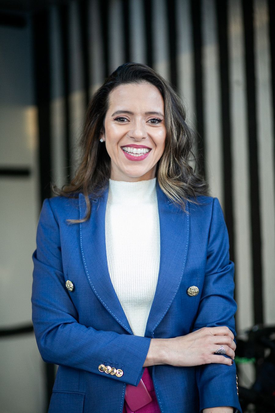 Mulher sorrindo, vestindo um blazer azul e com os braços cruzados, contra um fundo listrado escuro.