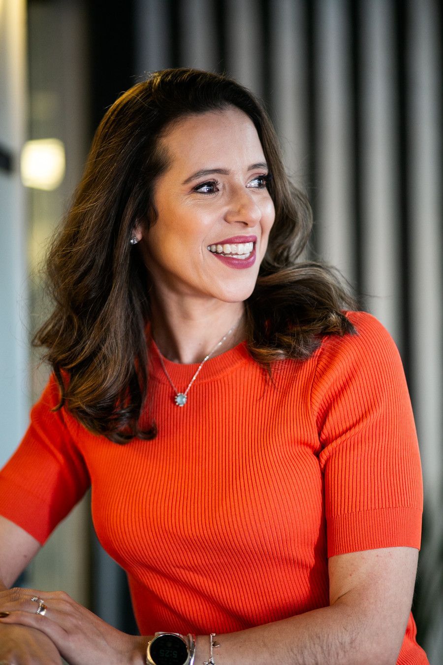Pessoa sorridente vestindo uma blusa laranja vibrante, sentada em um ambiente interno contra um fundo listrado com iluminação suave.