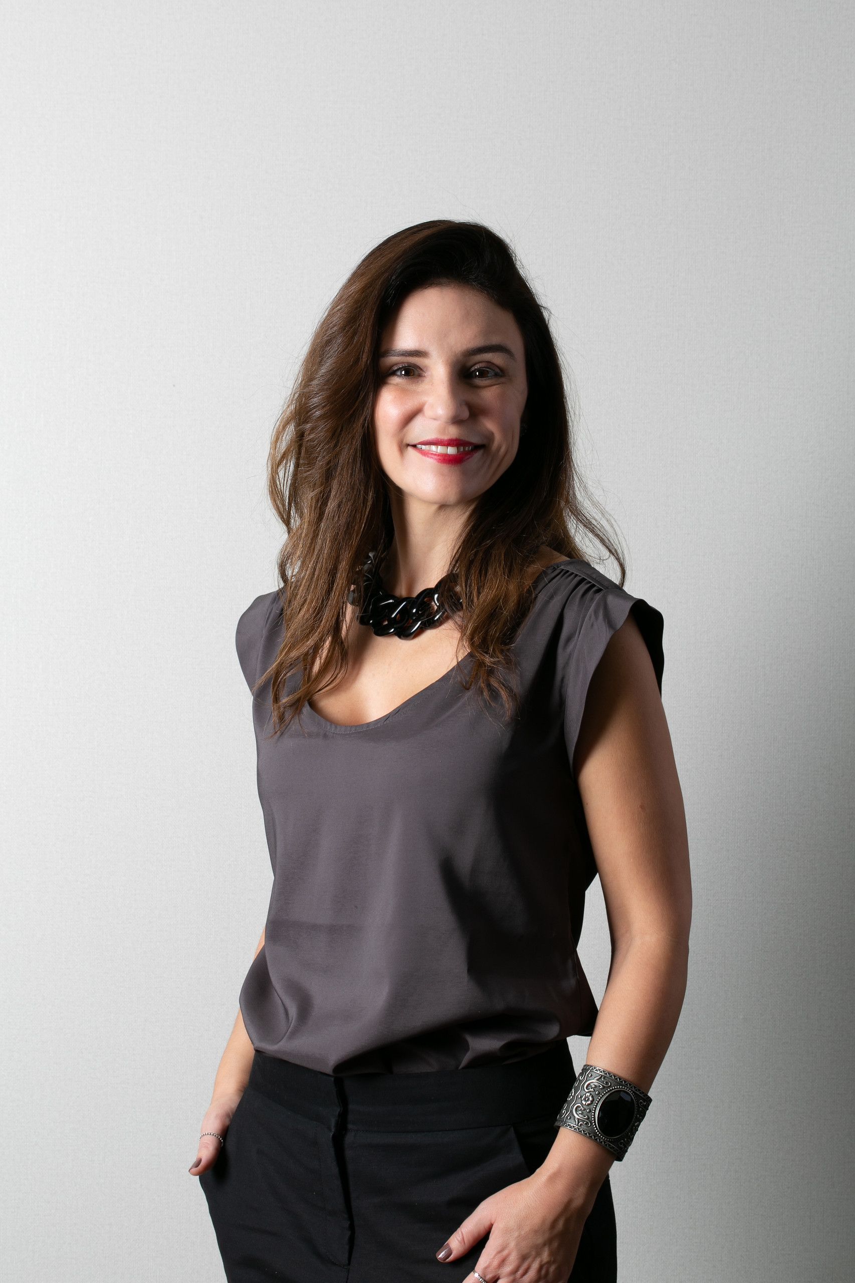 Mulher em pé contra uma parede clara, vestindo uma blusa cinza, calças pretas e pulseiras, com uma das mãos no bolso.