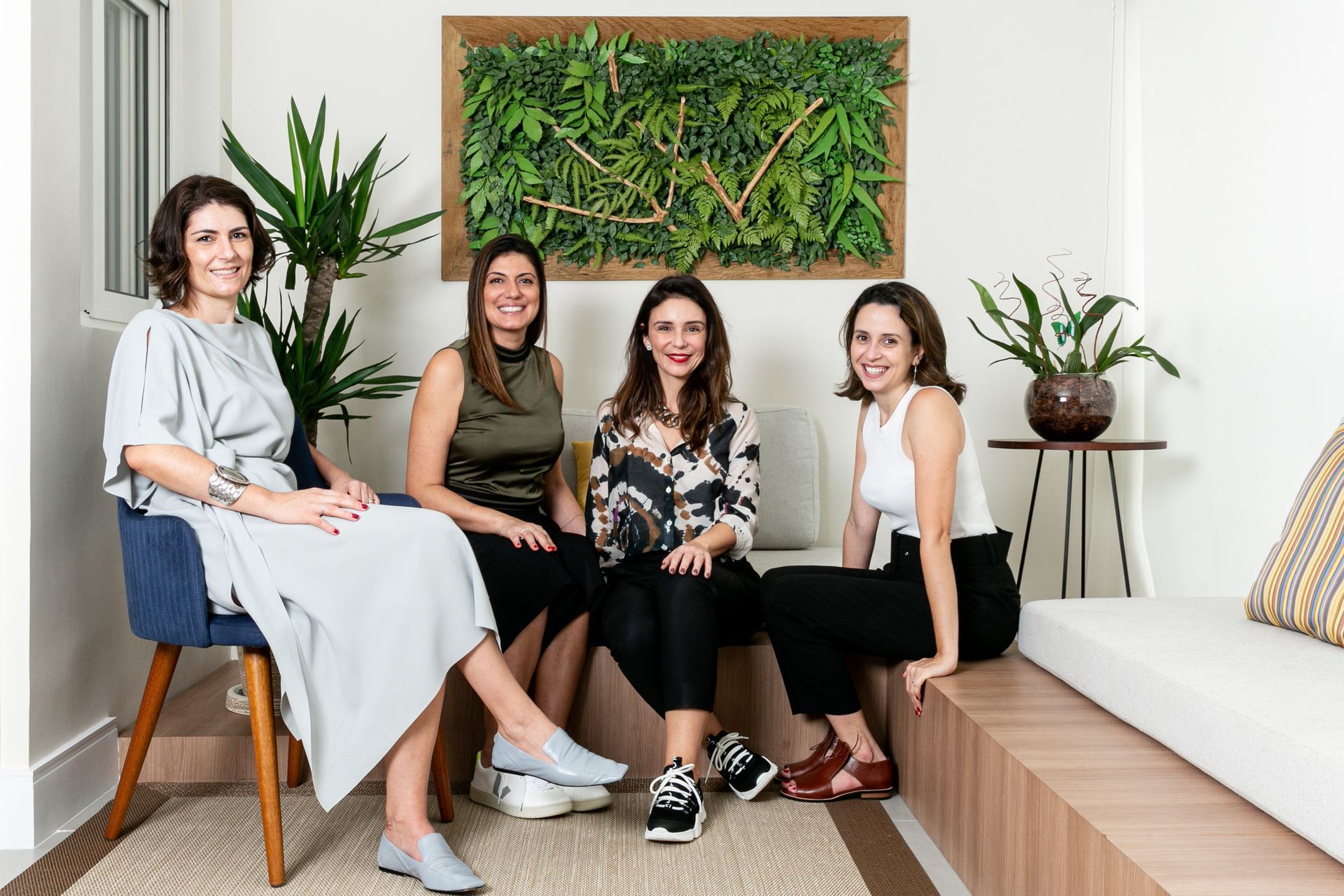 Quatro mulheres sentadas em uma sala de estar iluminada, com plantas e uma obra de arte verde na parede.