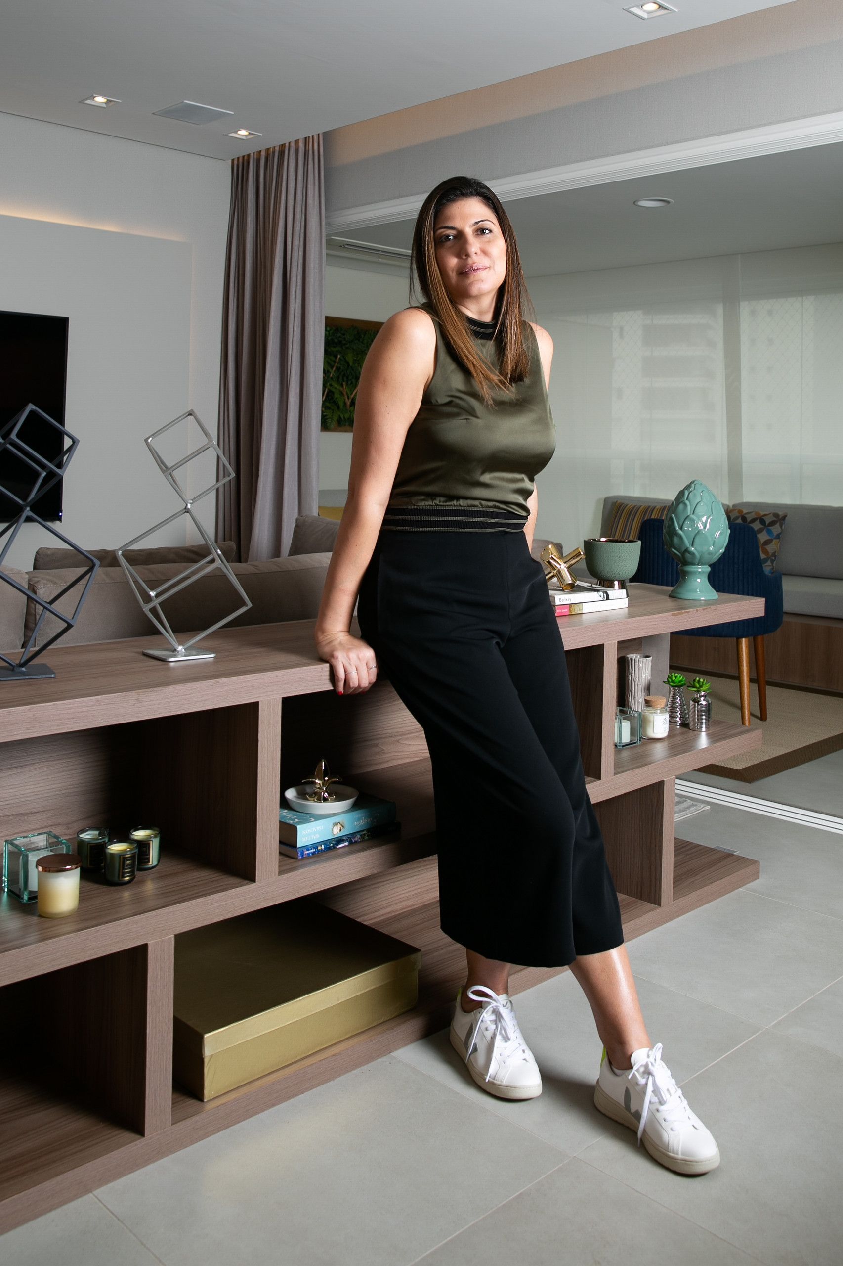 Mulher encostada em uma mesa em uma sala de estar moderna, vestindo uma blusa verde-oliva, calça preta e tênis brancos.