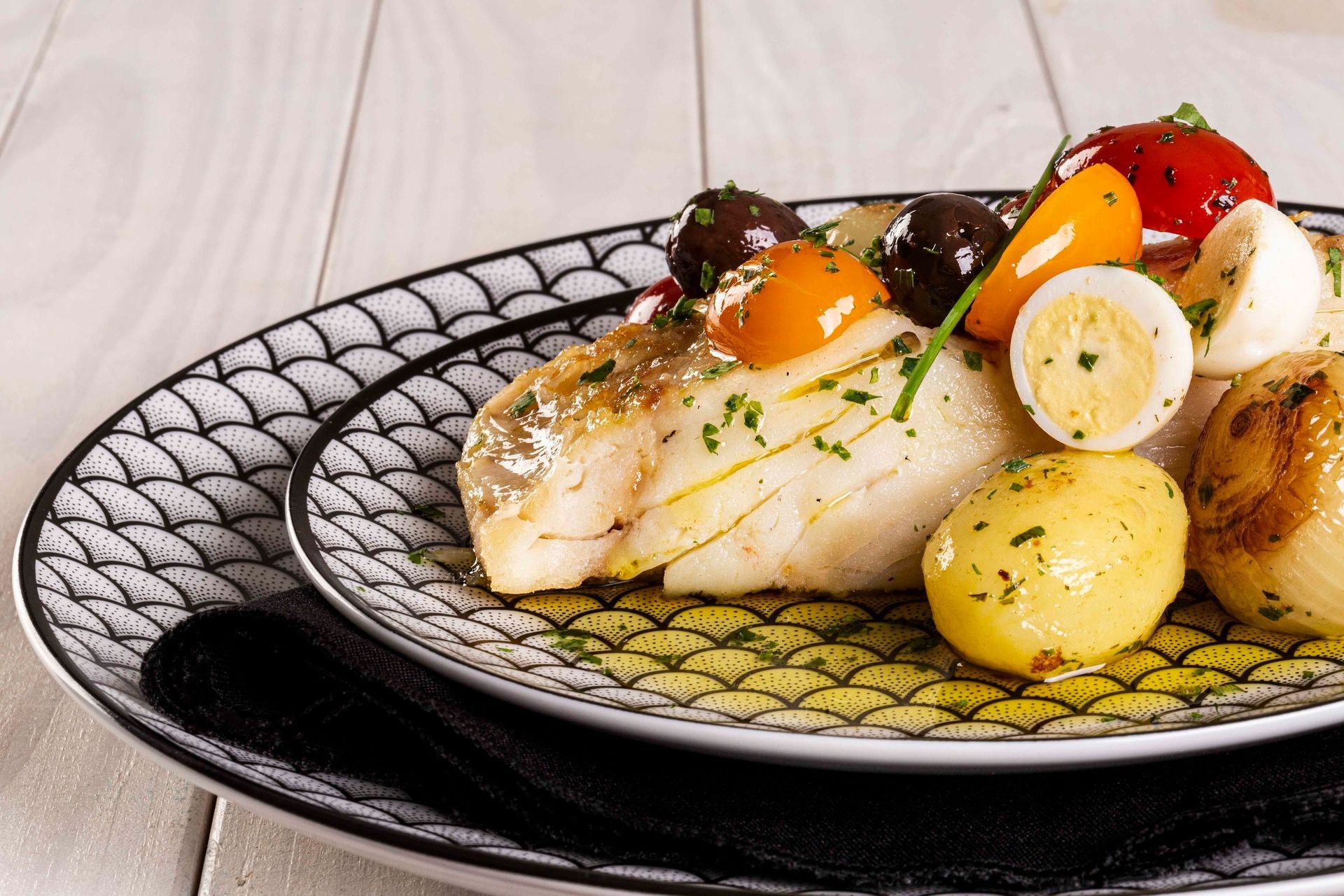 Peixe gourmet servido com batatas, tomates-cereja e ervas em um prato decorado.