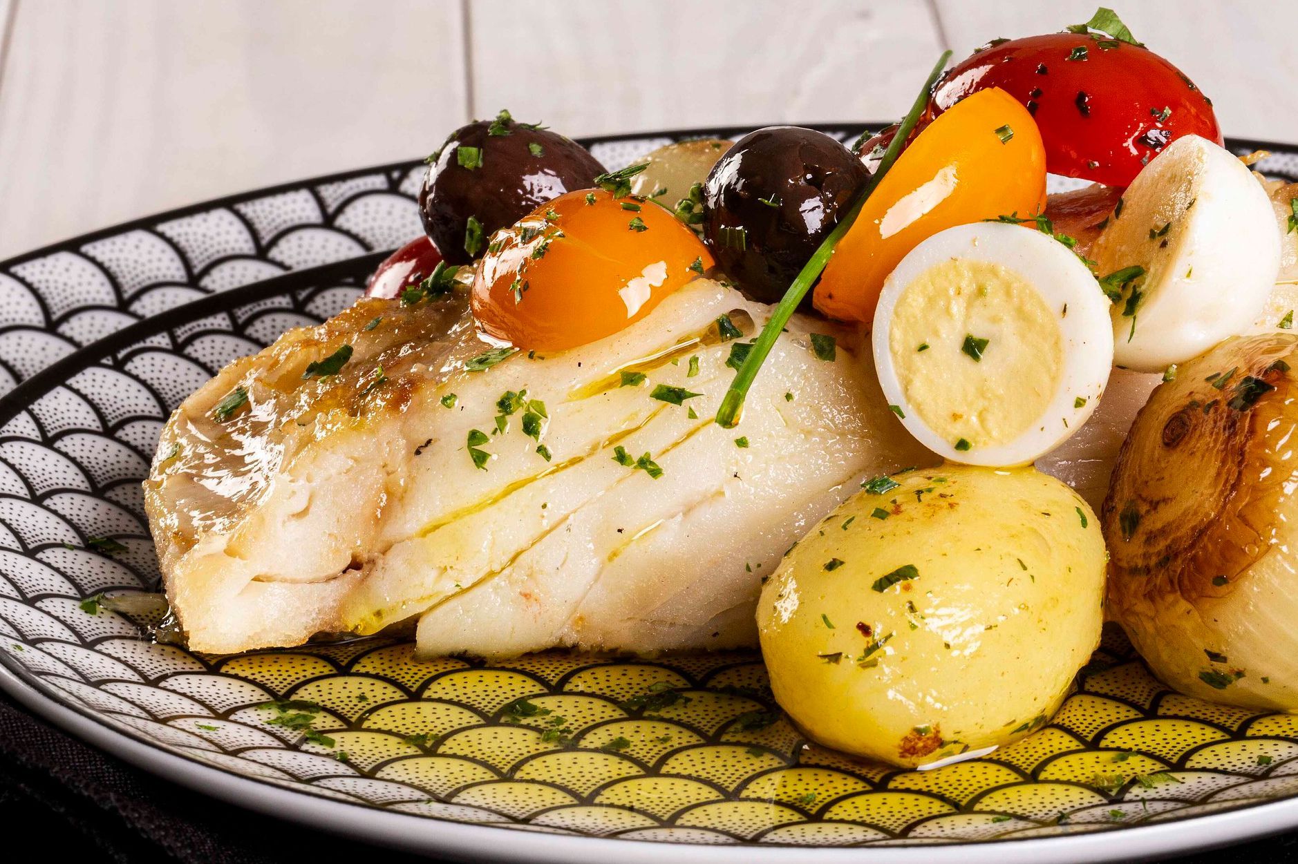 Filé de peixe servido com batatinhas, tomates-cereja e ervas em um prato decorativo com borda preta.