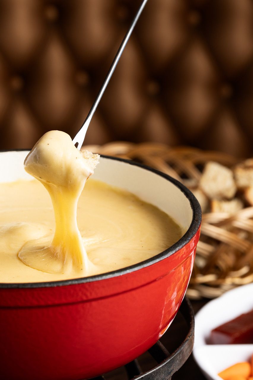 Fondue de queijo servido em uma panela vermelha, com pão sendo mergulhado em um espeto.