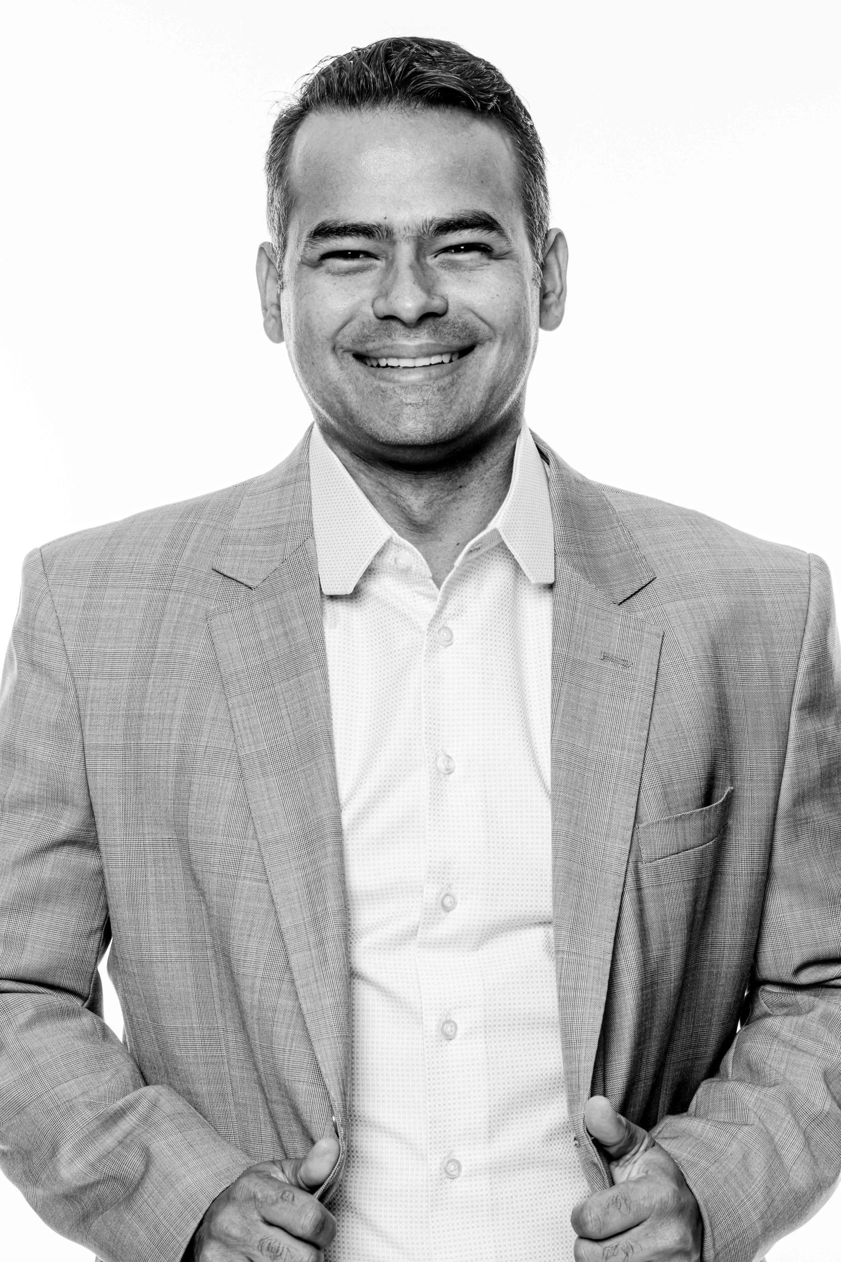Homem sorridente, vestindo um blazer claro e camisa branca, sentado contra um fundo branco liso.