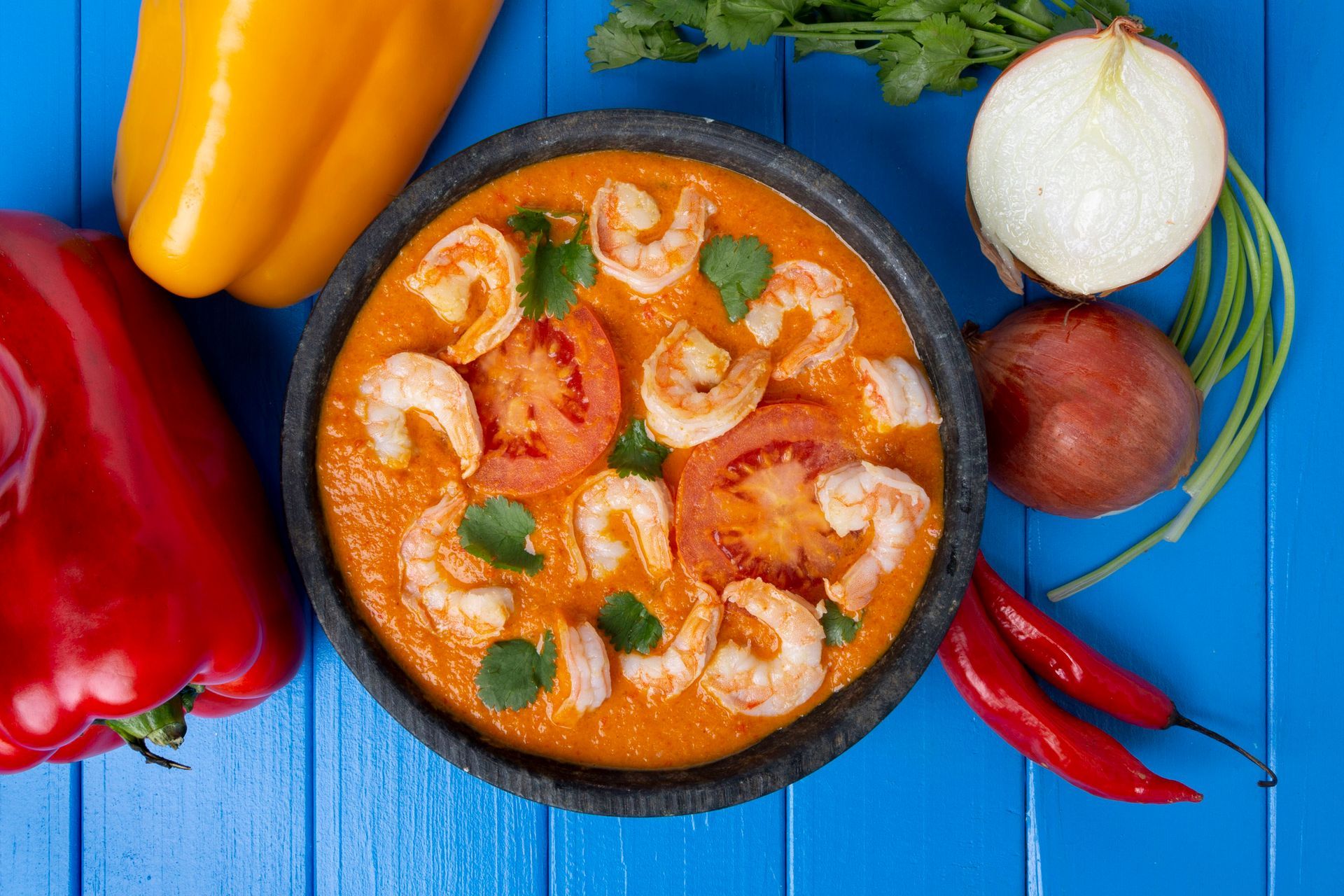 Curry de camarão em uma tigela com pimentões, cebola, tomates e pimentas sobre uma mesa de madeira azul.