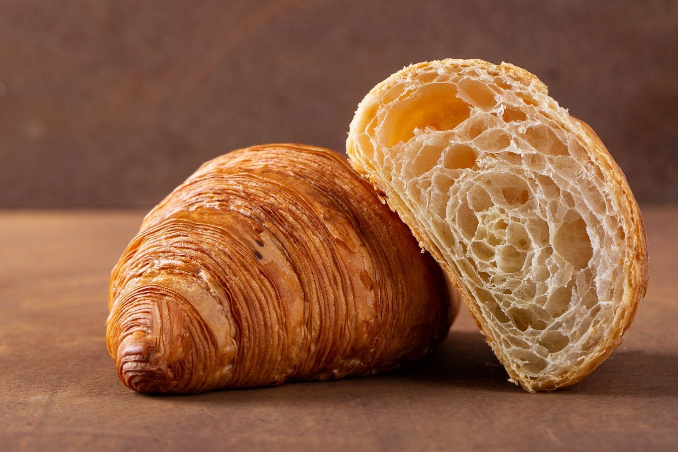 Dois croissants sobre uma mesa de madeira, um inteiro e outro aberto ao meio, revelando as camadas folhadas.