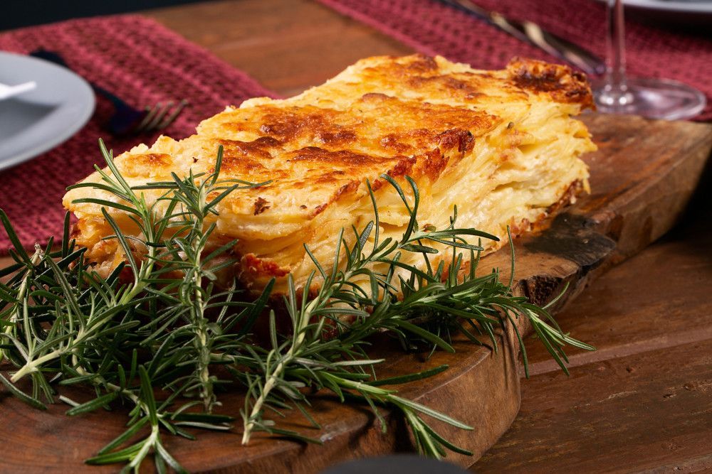 Caçarola gratinada com queijo e ervas, assada em uma tábua de madeira ao lado de uma taça de vinho e um prato.