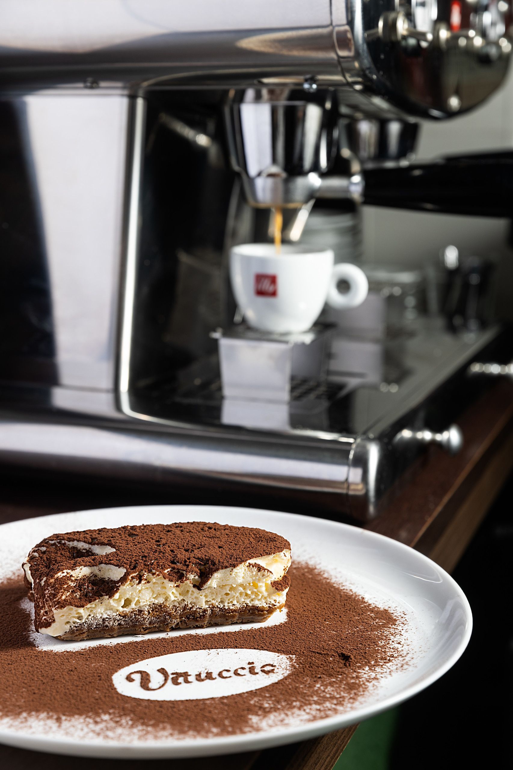 Máquina de café expresso servindo café em uma xícara branca ao lado de um prato de sobremesa no balcão de um café.
