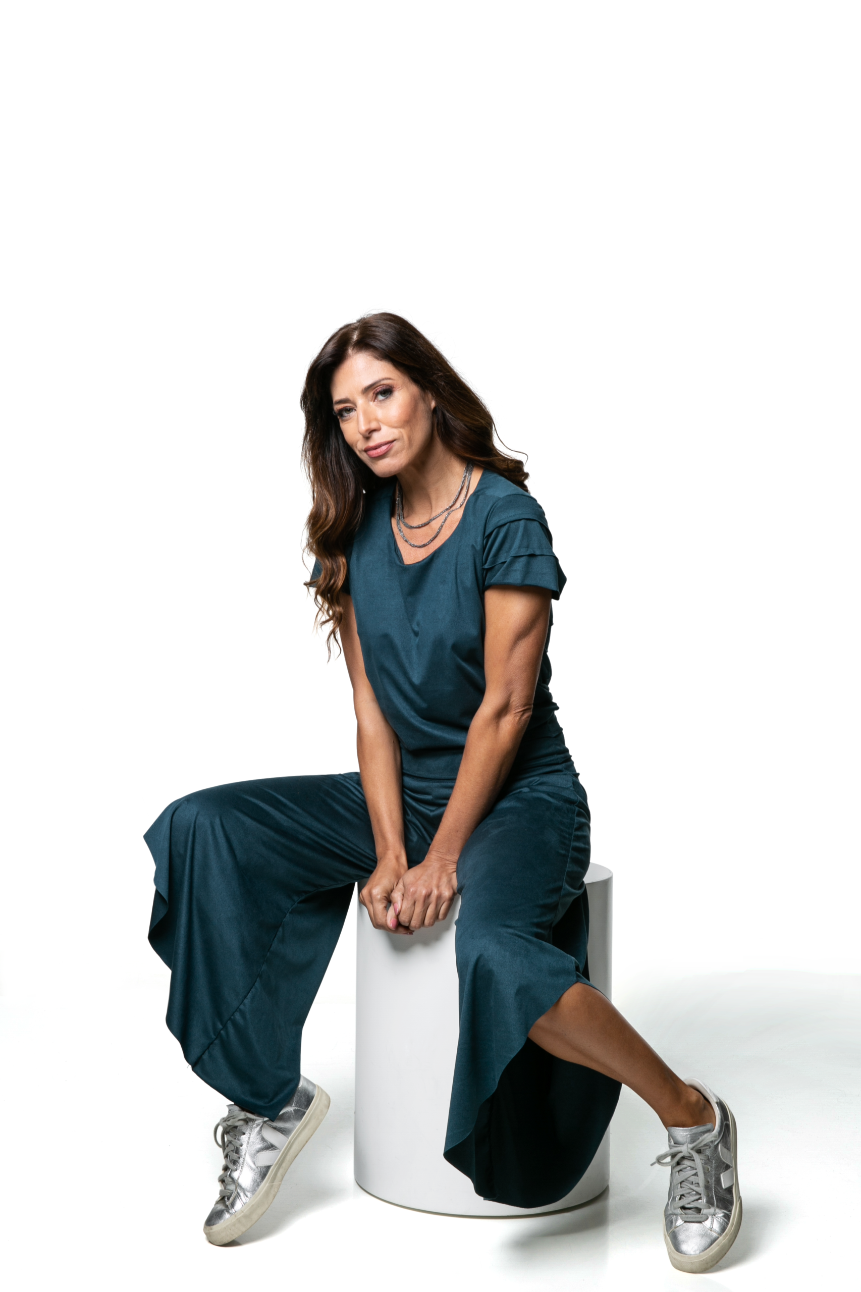 Mulher vestida com um conjunto verde-azulado, sentada em um cilindro branco contra um fundo branco, usando tênis prateados.