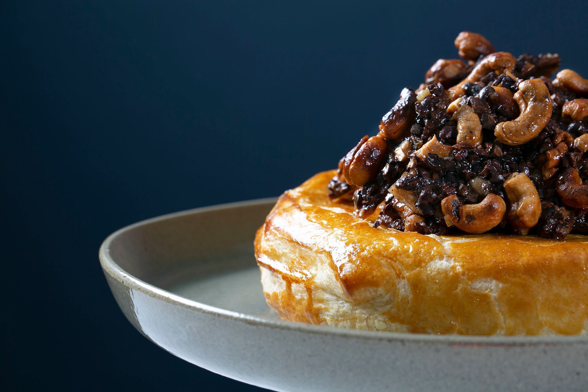Pastel coberto com nozes caramelizadas em um prato cinza sobre um fundo azul escuro.