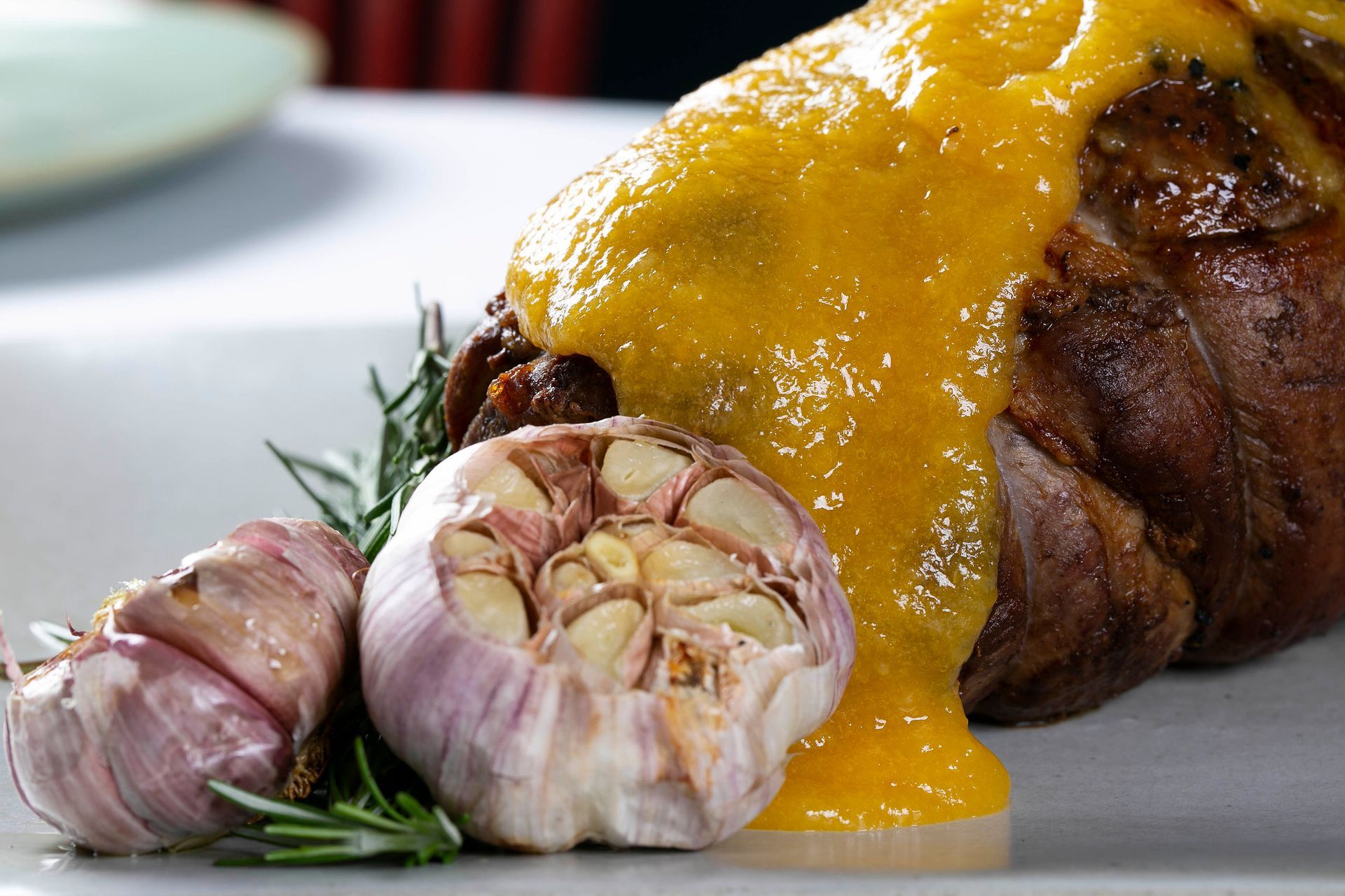Carne assada coberta com molho amarelo, com dentes de alho e alecrim em um prato branco.