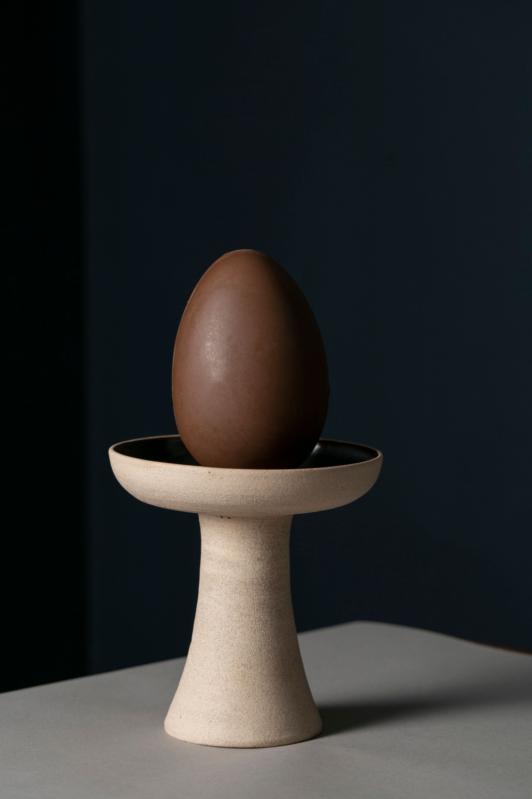 Um ovo marrom em pé dentro de uma tigela bege com pedestal, contra um fundo escuro.