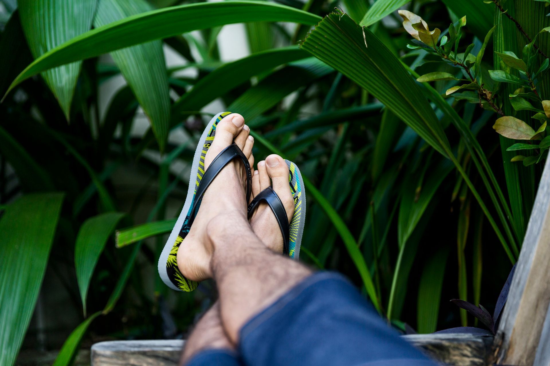 Pés de chinelo apoiados em um banco em meio a plantas tropicais verdejantes.