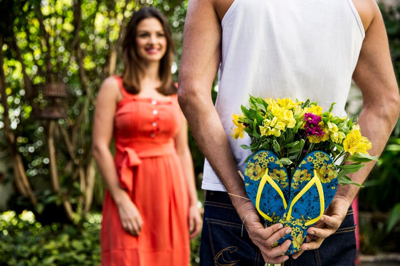 Uma pessoa segurando um buquê atrás das costas está de frente para uma mulher sorridente vestida com um vestido coral, ao ar livre.